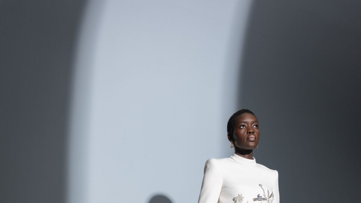 Portrait of a woman in a white embroidered top looking upward against a gray wall with a light streak on the left side.