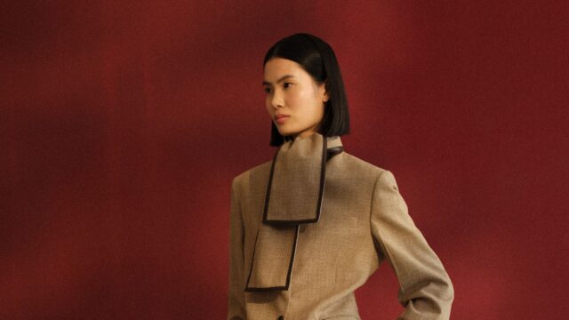 Woman with a short black bob wearing a beige coat with dark trim, posed against a red wall.