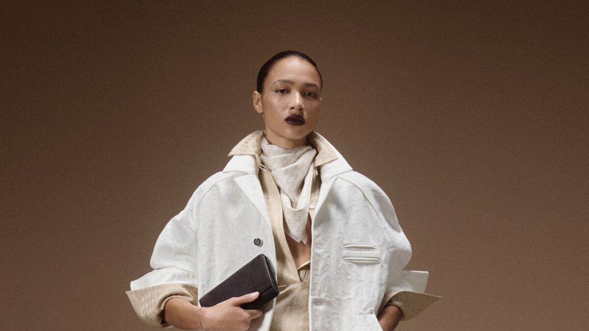 Fashion model in a light beige trench coat holding a black clutch against a warm brown studio backdrop.