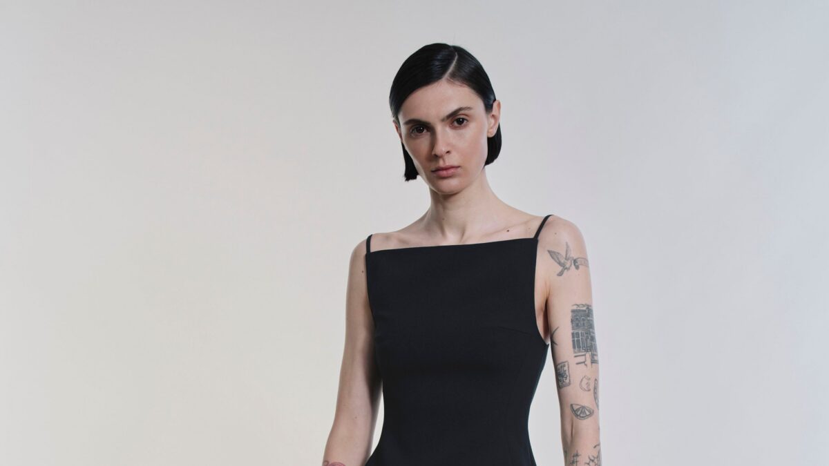 Woman with short black hair wearing a black sleeveless dress, standing against a pale gradient wall with visible tattoos on her left arm.