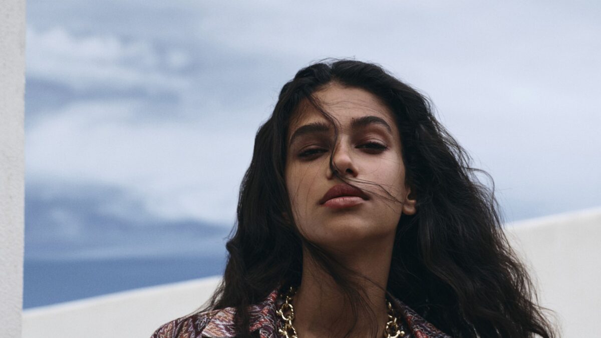 Close-up portrait of a woman with dark hair outdoors, blue sky behind her, wearing a patterned top and gold necklace.
