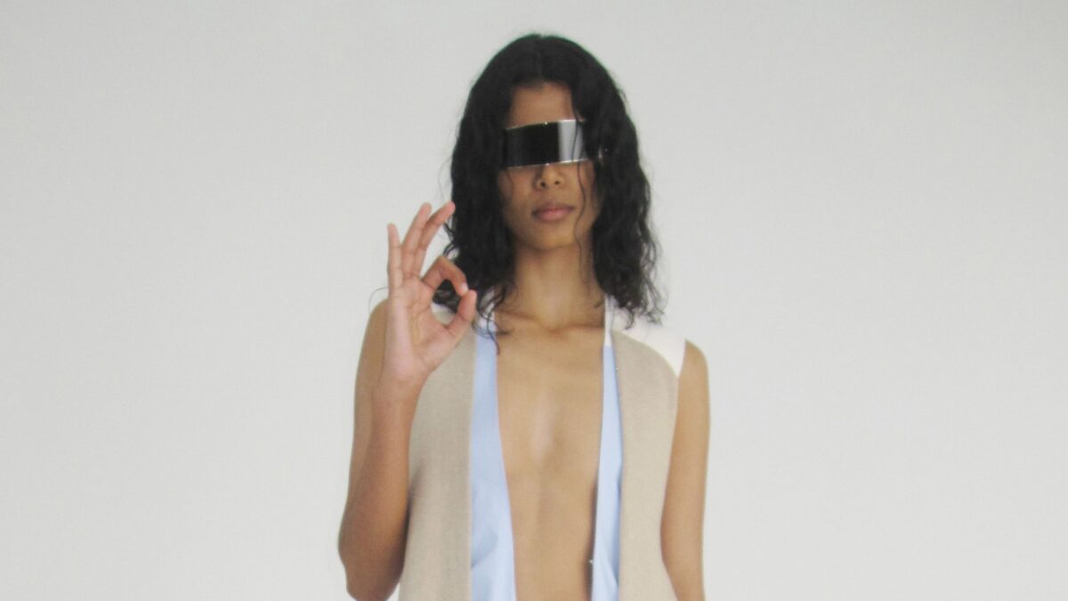 Person with long dark hair wearing a futuristic rectangular visor and making an OK sign with their right hand, against a plain light background.