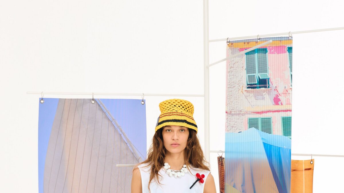 Model wearing a yellow straw hat and white sleeveless top stands in front of colorful fashion banners.