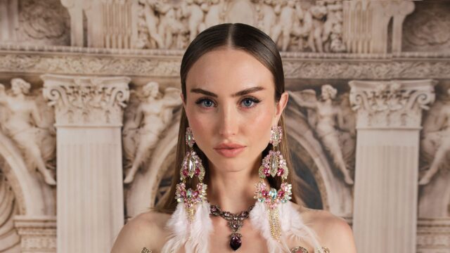 Close-up of a woman with blue eyes wearing elaborate pink crystal jewelry against a marble relief backdrop.