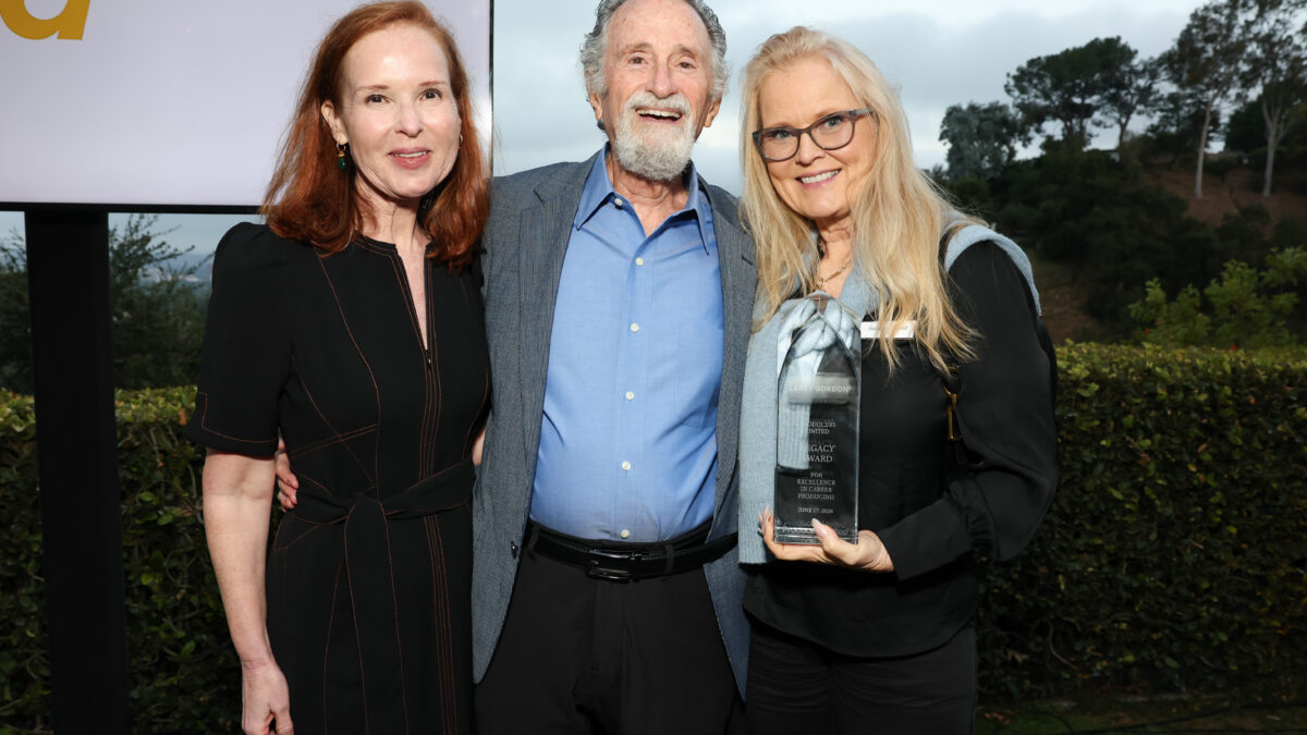 Three adults posing outdoors: a man with gray hair and beard in the center holding a tall award, flanked by two smiling women on either side.