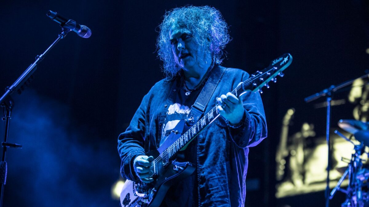 Guitarist with curly hair in a denim jacket plays a black electric guitar on a blue-lit stage.