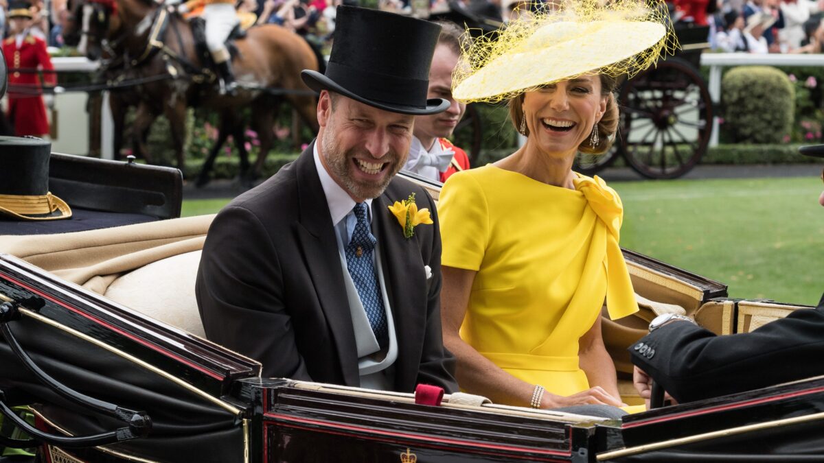 Smiling couple in a black-and-gold horse-drawn carriage; man in a tux with a top hat and yellow boutonniere, woman in a bright yellow dress and elaborate yellow hat; spectators blurred in background.