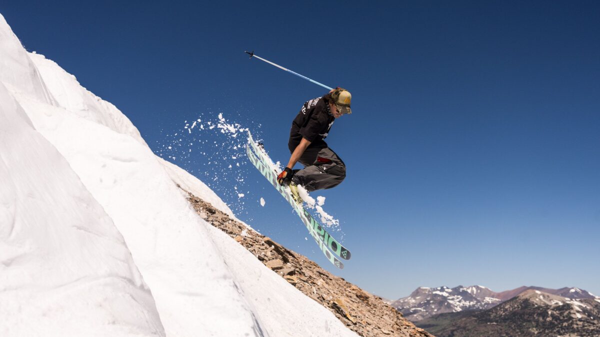 Skier in mid-air performing a jump off a snow-covered ridge with a spray of snow against a clear blue sky.
