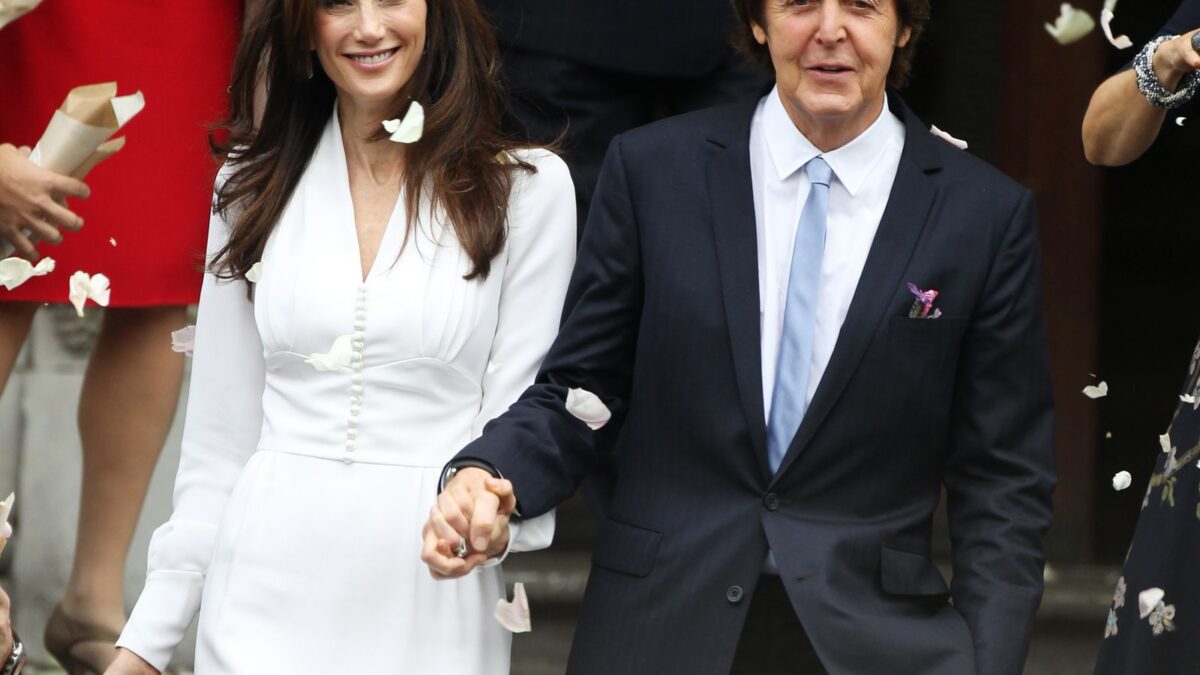 Couple holding hands after a ceremony, woman in white dress and man in dark suit, petals falling around them.
