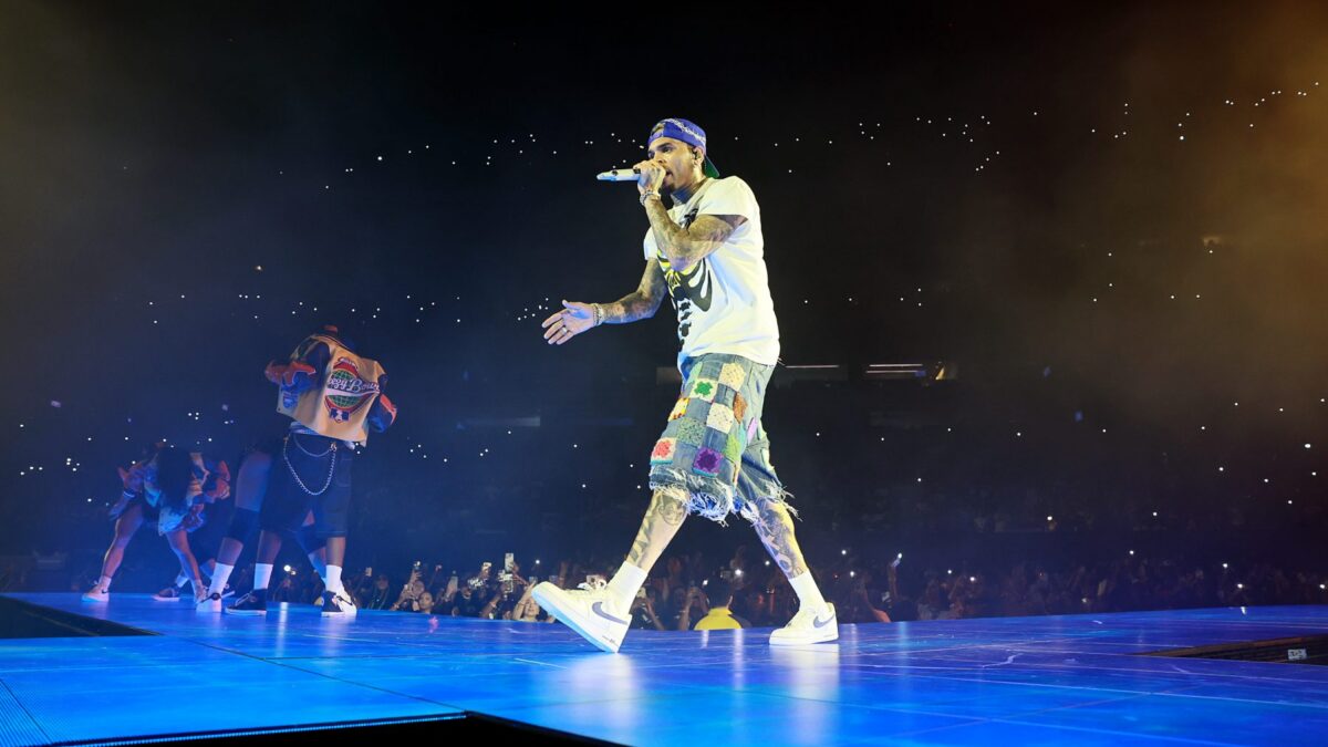 Male rapper wearing a bandana and patchwork shorts performs center stage, mic in hand, blue-lit floor and cheering crowd with phone lights in a night concert.
