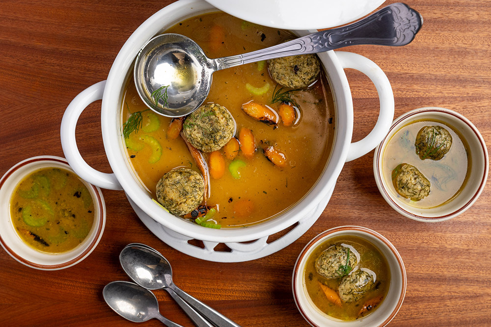 White pot of hearty vegetable soup with meatballs and carrot pieces on a wooden table, surrounded by small bowls and spoons.