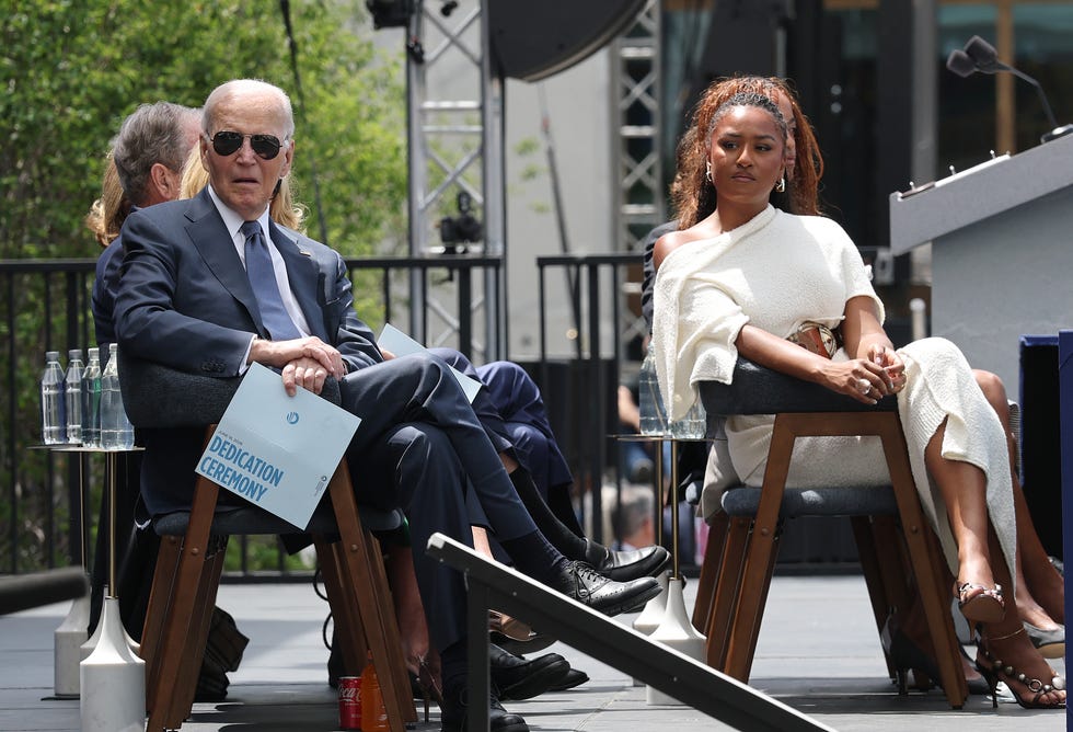 Two formally dressed attendees sit on stage at an outdoor dedication ceremony; the man holds a program reading 'Dedication Ceremony'.