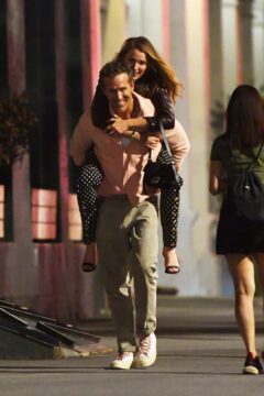 Smiling couple on a city sidewalk at night; the woman rides piggyback on the man’s back, both casually dressed.