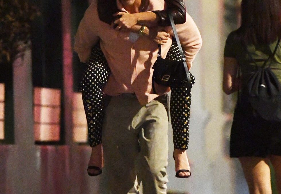 Smiling couple on a city sidewalk at night; the woman rides piggyback on the man’s back, both casually dressed.