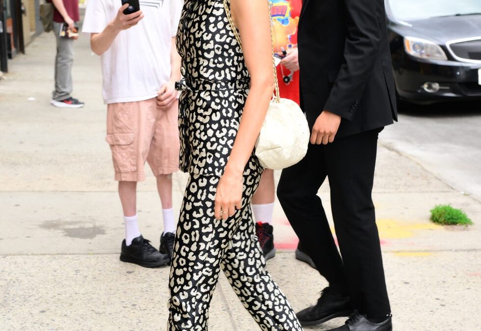 Woman in a black-and-white leopard print jumpsuit and black strappy heels walks on a city sidewalk with a man in a suit behind her, onlookers in the background.