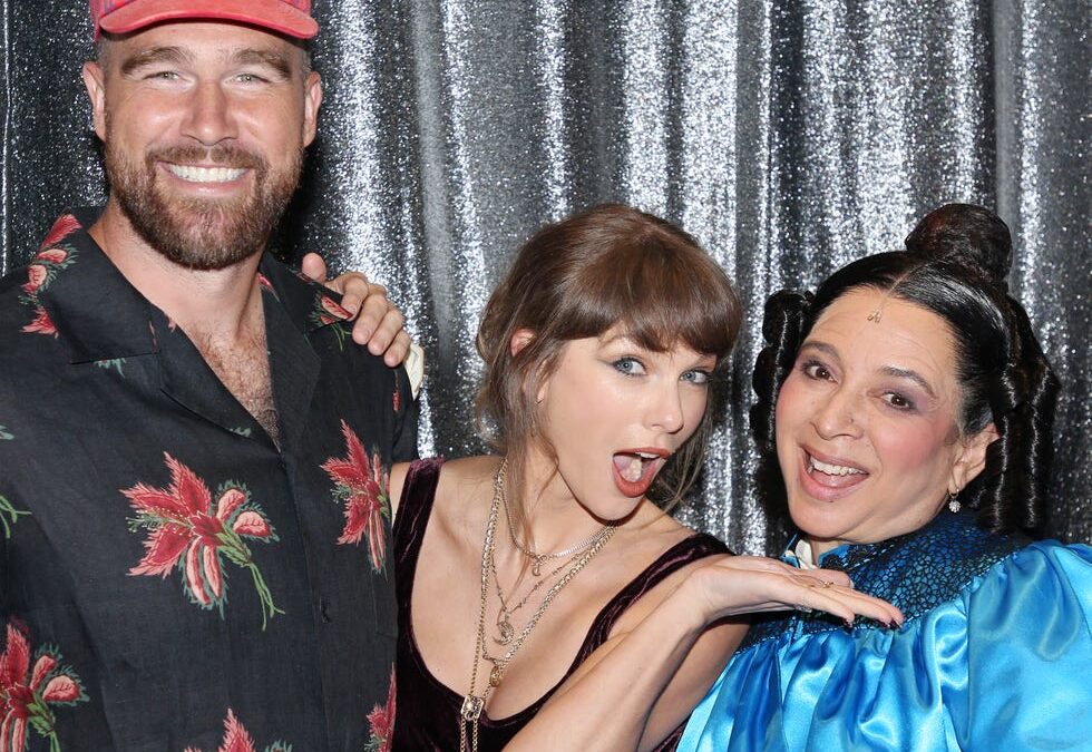 Three people posing together in front of a glittery backdrop; man on the left wears a red cap and floral shirt, woman in the middle wears a burgundy dress, woman on the right wears a blue outfit with ruffles.
