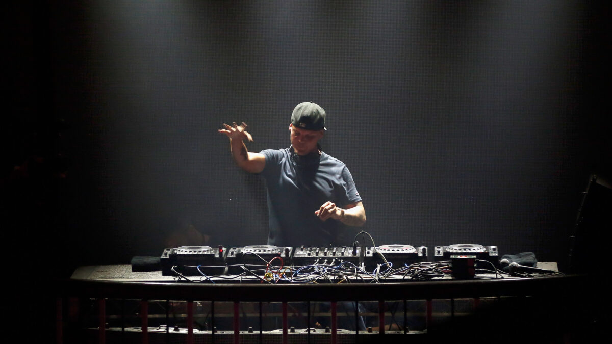 DJ wearing a cap performing behind a crowded mixer setup on a dimly lit stage with dark backdrop