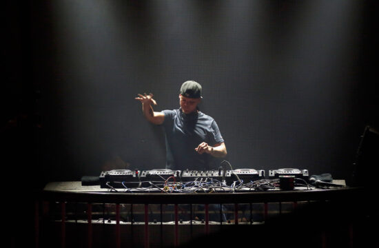 DJ wearing a cap performing behind a crowded mixer setup on a dimly lit stage with dark backdrop