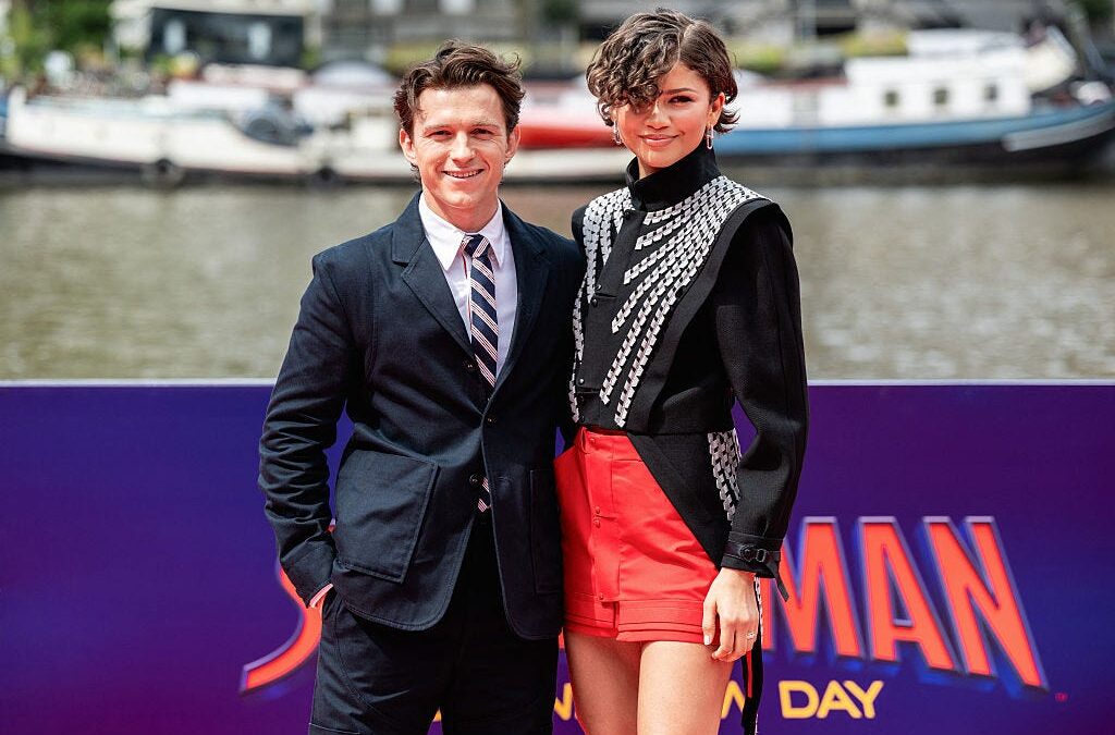 Two people pose for photos at an event by a water backdrop; man in a navy suit and striped tie, woman in a black embellished top and red shorts, in front of a purple banner reading 'SPIDERMAN DAY'.