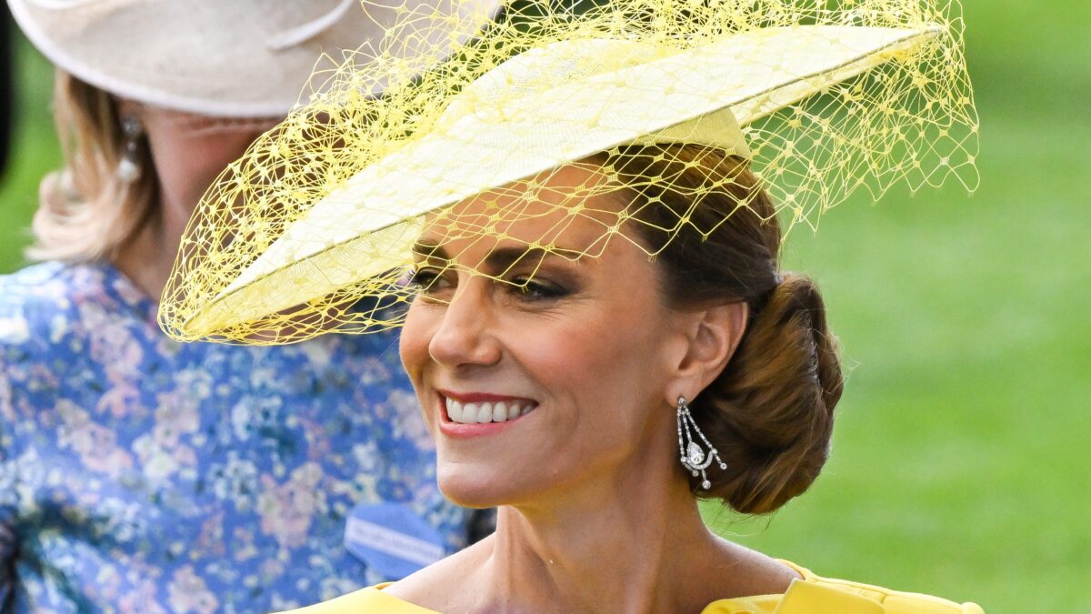 Smiling woman in a bright yellow dress wearing a decorative yellow netted fascinator outdoors.