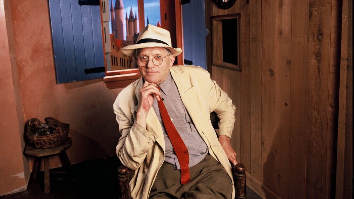 Older man in a beige suit, red tie and fedora sits in a rustic studio with a painted city backdrop.