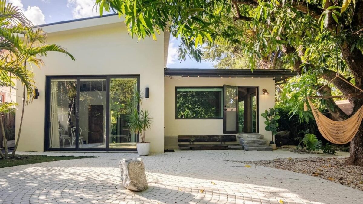 Modern single-story house with large glass sliding doors, light exterior, and a shaded cobblestone courtyard
