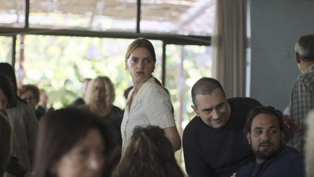A group of people at a casual indoor gathering; a woman in a white blouse looks to the side while two men relax and chat nearby.