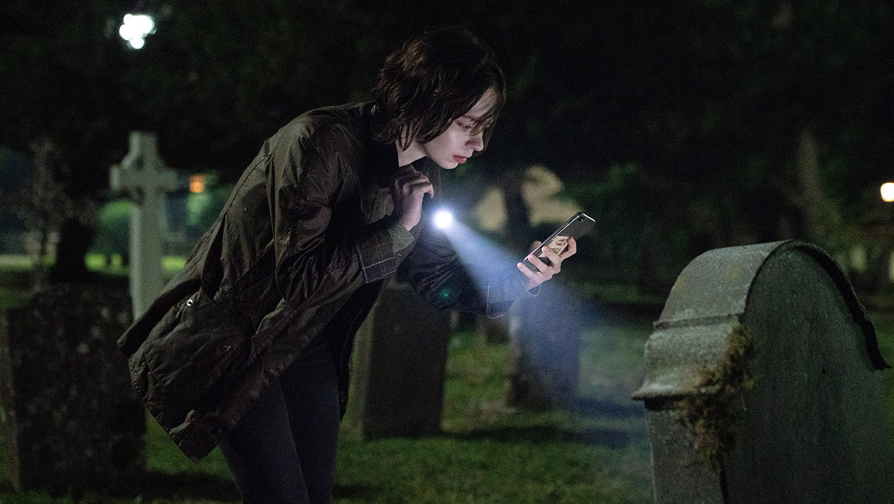 "Person in dark clothing examines a gravestone at night in a cemetery, using the flashlight on their phone to illuminate the scene; the background is dimly lit with other gravestones and trees."