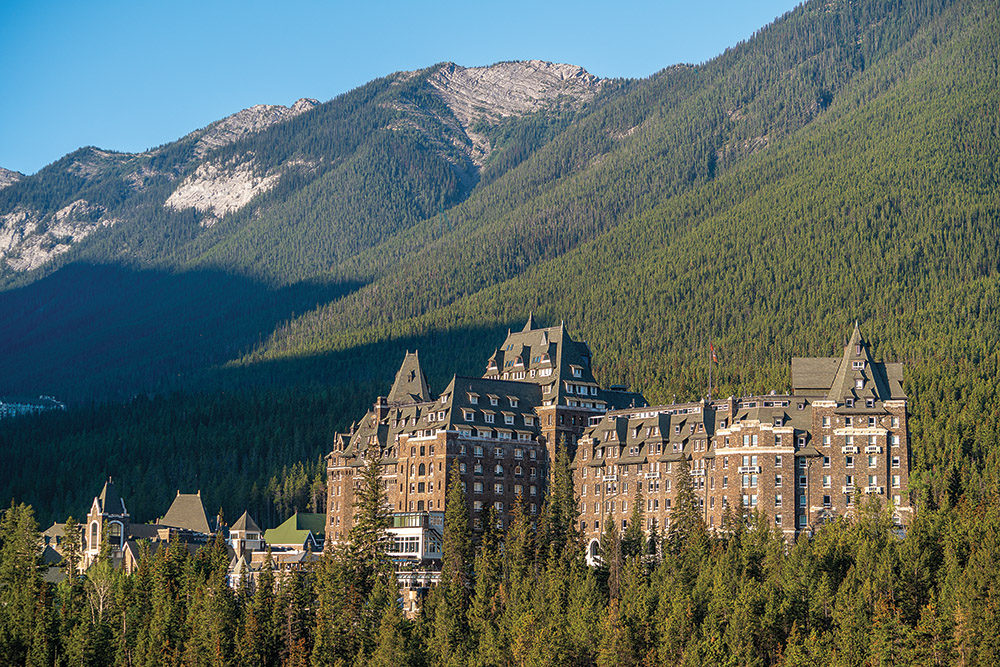 Castle-like stone hotel nestled in a dense evergreen forest with mountain peaks in the background.