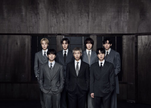Group of men in gray suits posing in a doorway against a dark, industrial wall backdrop.