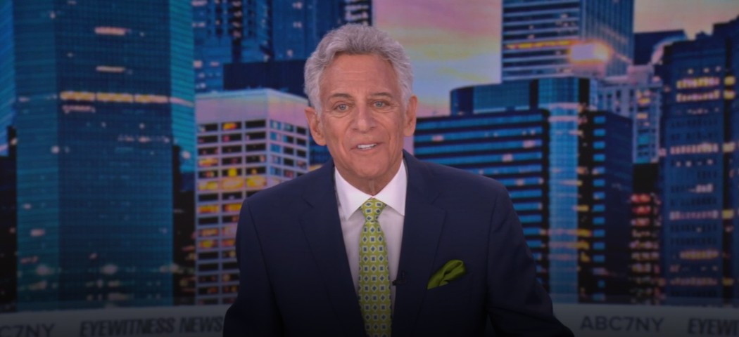 Male TV news anchor in a dark suit, white shirt, and green patterned tie sits at the desk with a city skyline backdrop behind him.