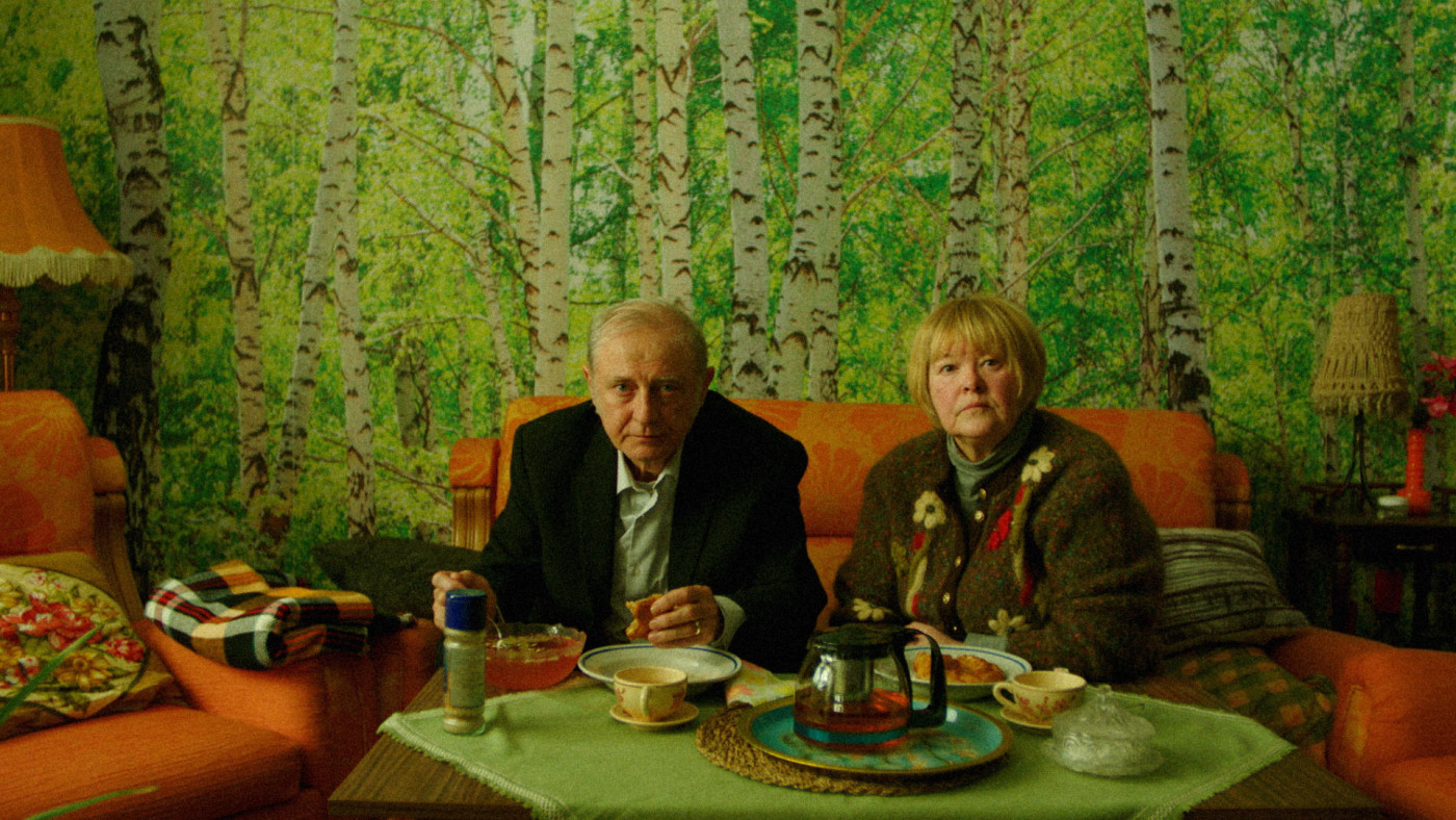 An elderly couple sits at a table with tea and snacks on an orange sofa in a living room with forest-pattern wallpaper behind them far wall.