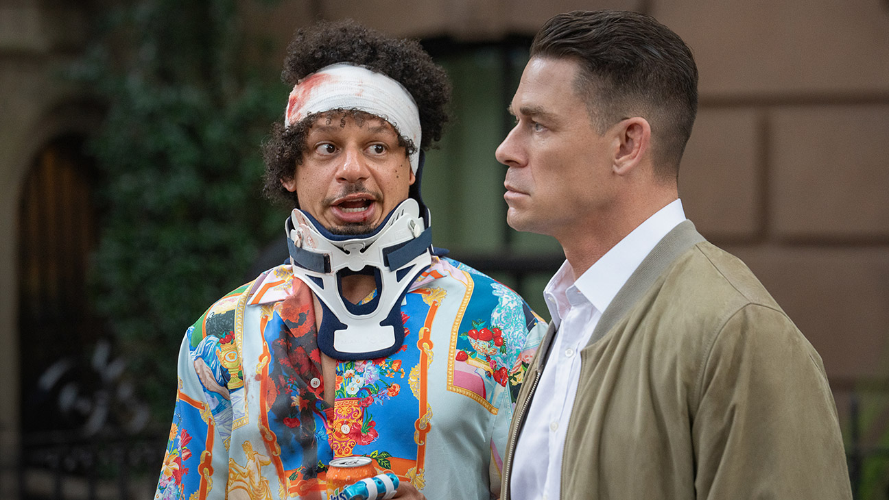 "Two men outdoors: one wears a colorful shirt, head bandage, and neck brace, holding a can, looking surprised; the other in a tan jacket and white shirt, looking serious. Urban background with greenery and brownstone building."