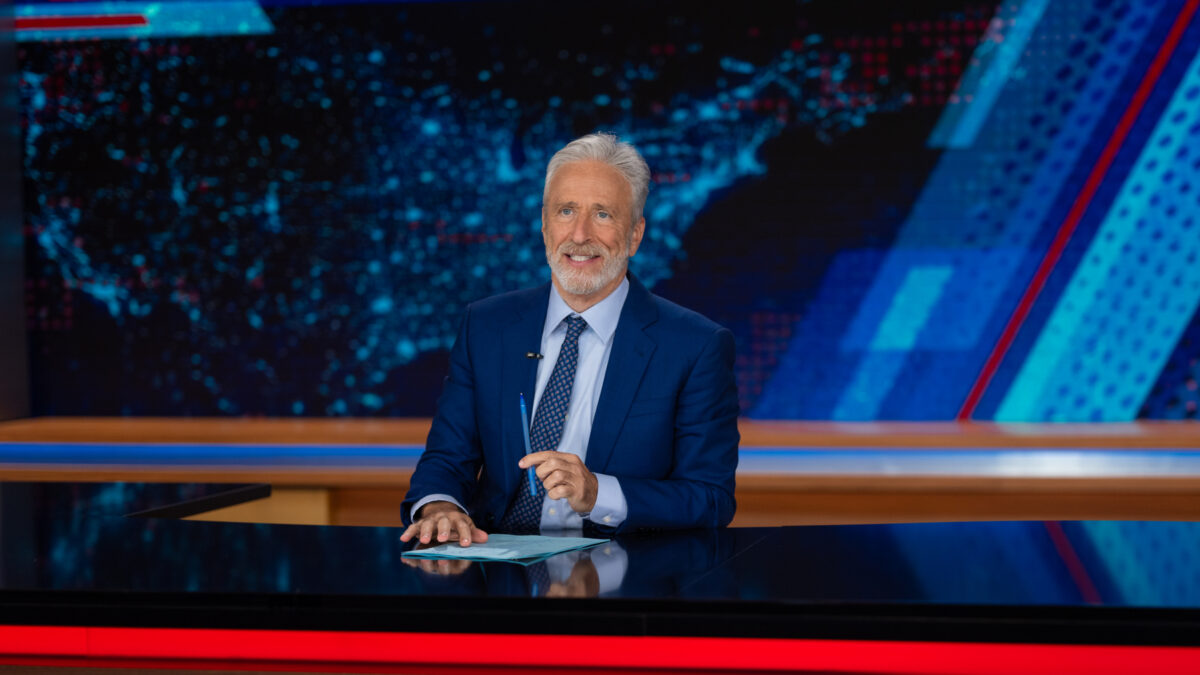 News anchor in a navy suit sits at a desk with papers and a pen, against a blue/red high-tech studio background.