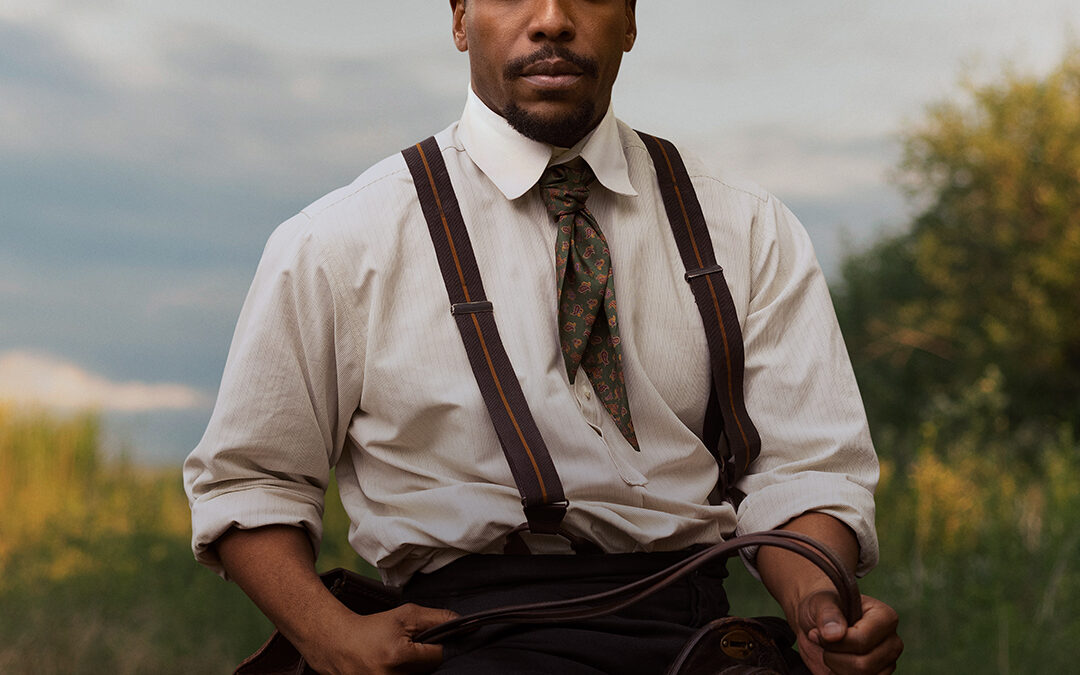 Man in a black fedora, white shirt with dark suspenders and a green patterned tie, riding a brown horse outdoors.