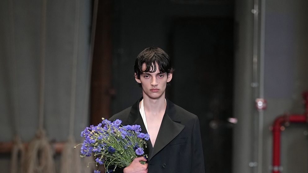 Male model in a black blazer backstage, holding a bouquet of purple flowers dimly lit hallway