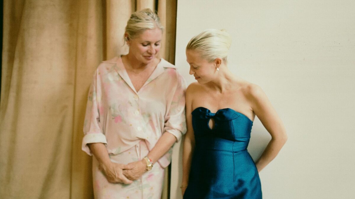 Two women standing close together by a beige curtain; one wears a pink floral robe, the other a teal dress, both looking down.
