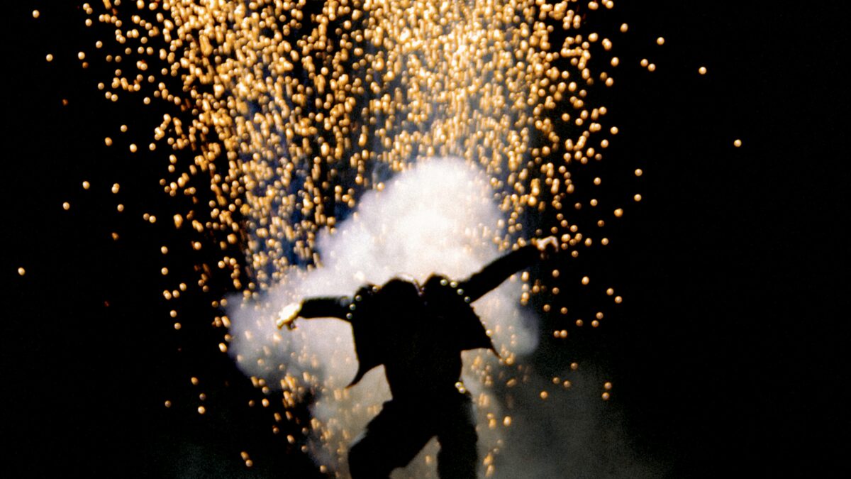 Silhouetted performer jumping on stage as orange sparks explode overhead against a dark background.