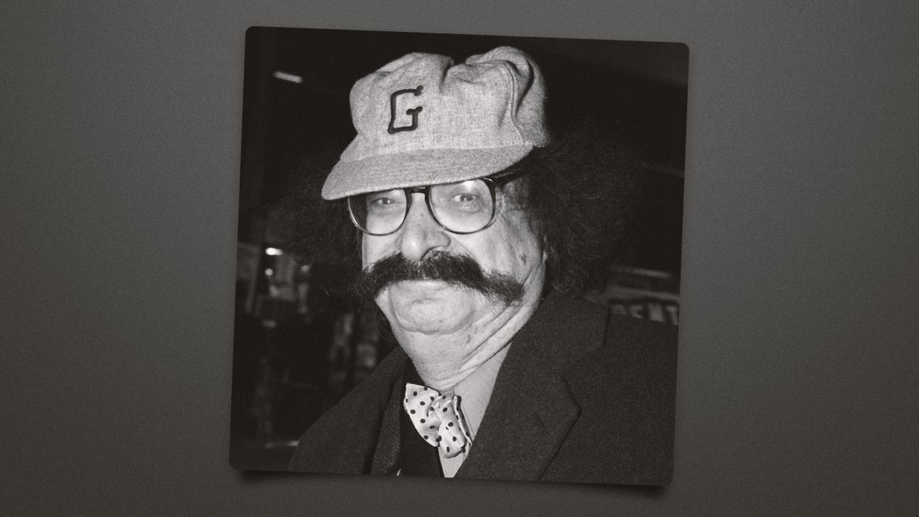 Black-and-white portrait of a man with a large mustache, round glasses, and a cap with the letter G, dressed in a blazer and polka-dot bow tie.