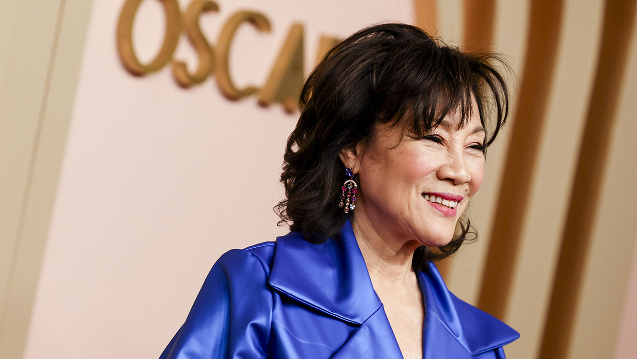 Smiling woman in a bright blue blazer posing at a red-carpet event with an Oscars backdrop behind her.