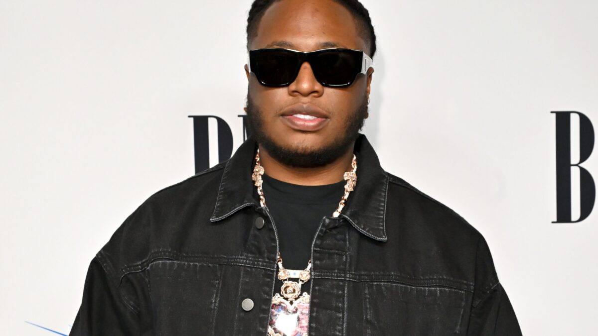 Man wearing oversized black sunglasses, black denim jacket, and a gold chain posing for a photo against a white backdrop at a fashion event.
