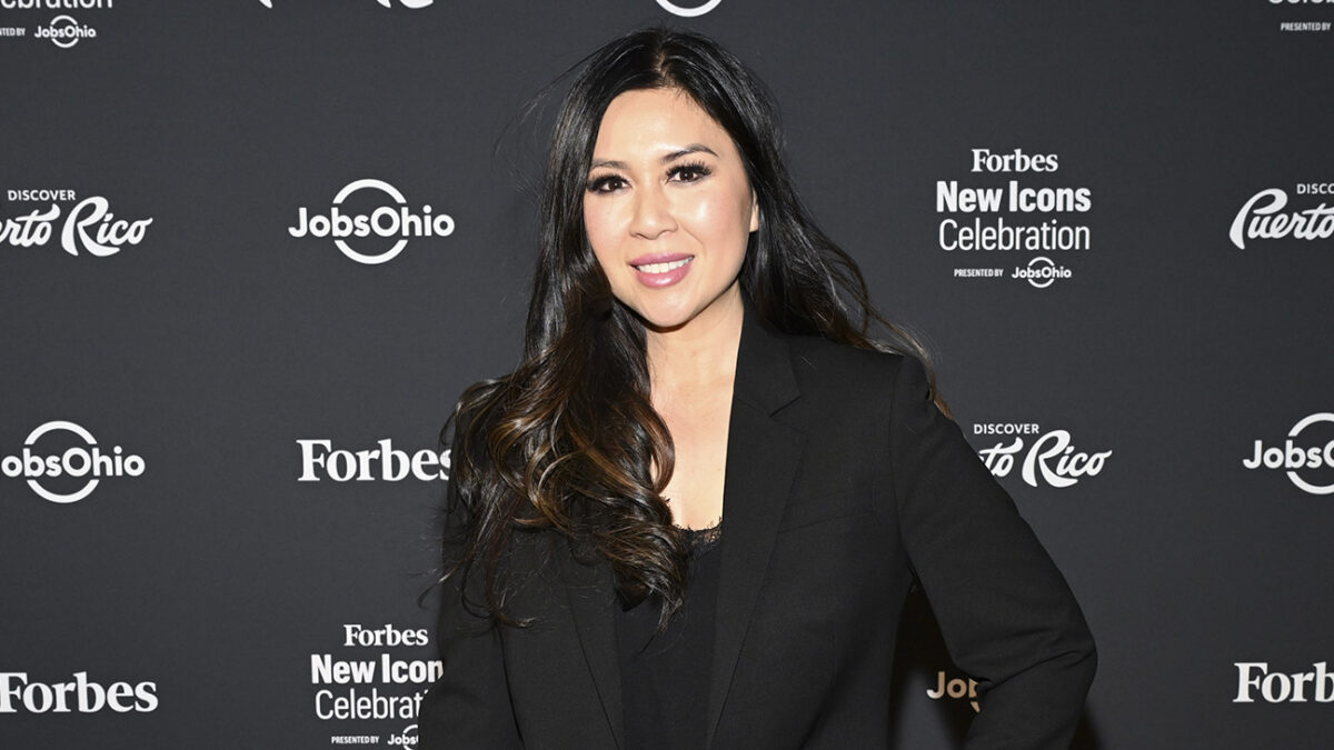 Woman with long dark hair in a black blazer posing on a dark backdrop at Forbes New Icons Celebration.