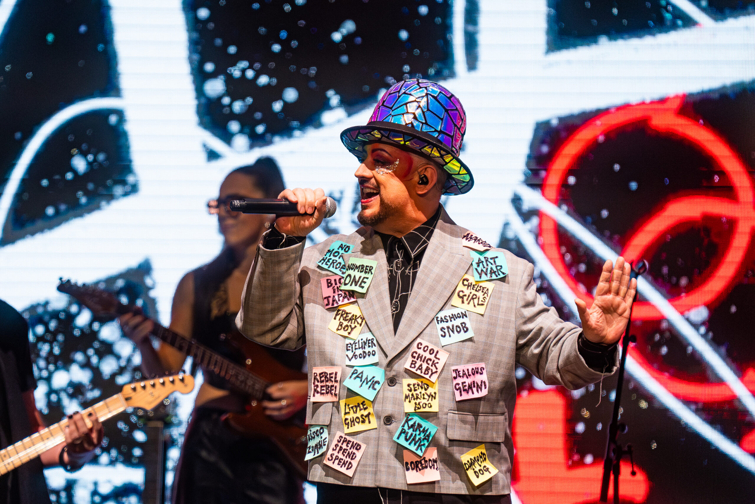 Singer on stage wearing a colorful mosaic hat and a blazer full of sticky notes, singing into a microphone.