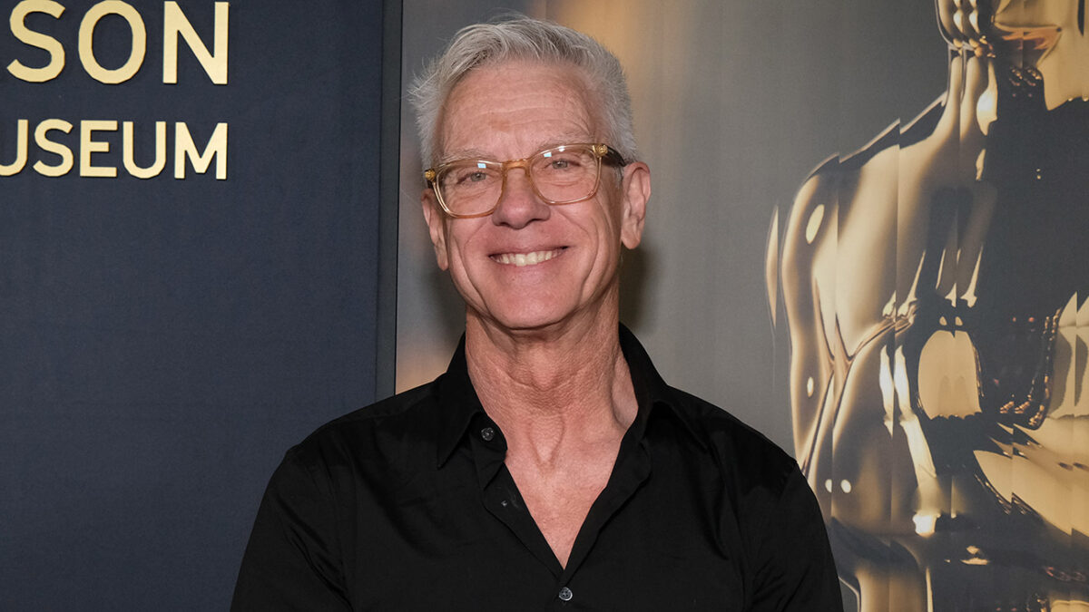 Smiling older man with gray hair and glasses posing in front of a museum backdrop.