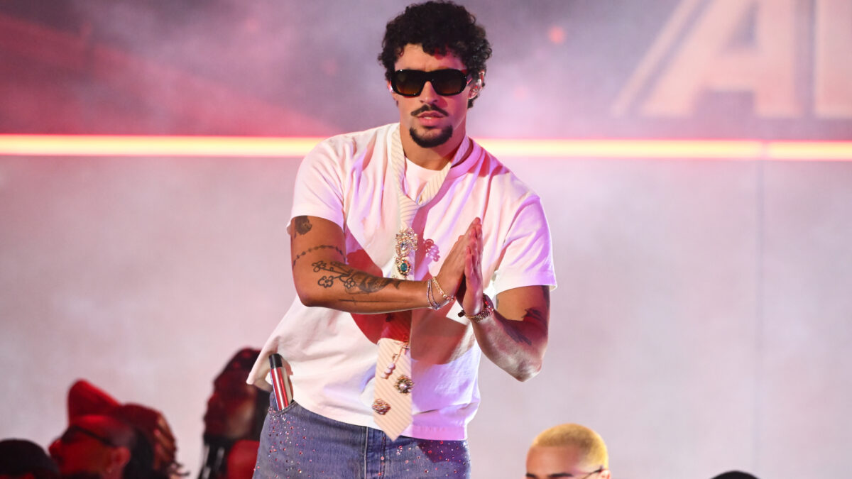 Male performer wearing sunglasses and a white T-shirt on stage, hands pressed together in a prayer-like pose, colorful lights and smoke in background.