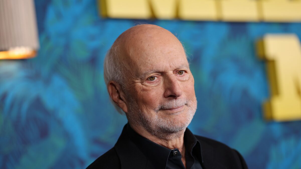 Portrait of an older man with a white beard, wearing a black shirt, against a blue and yellow mural background.
