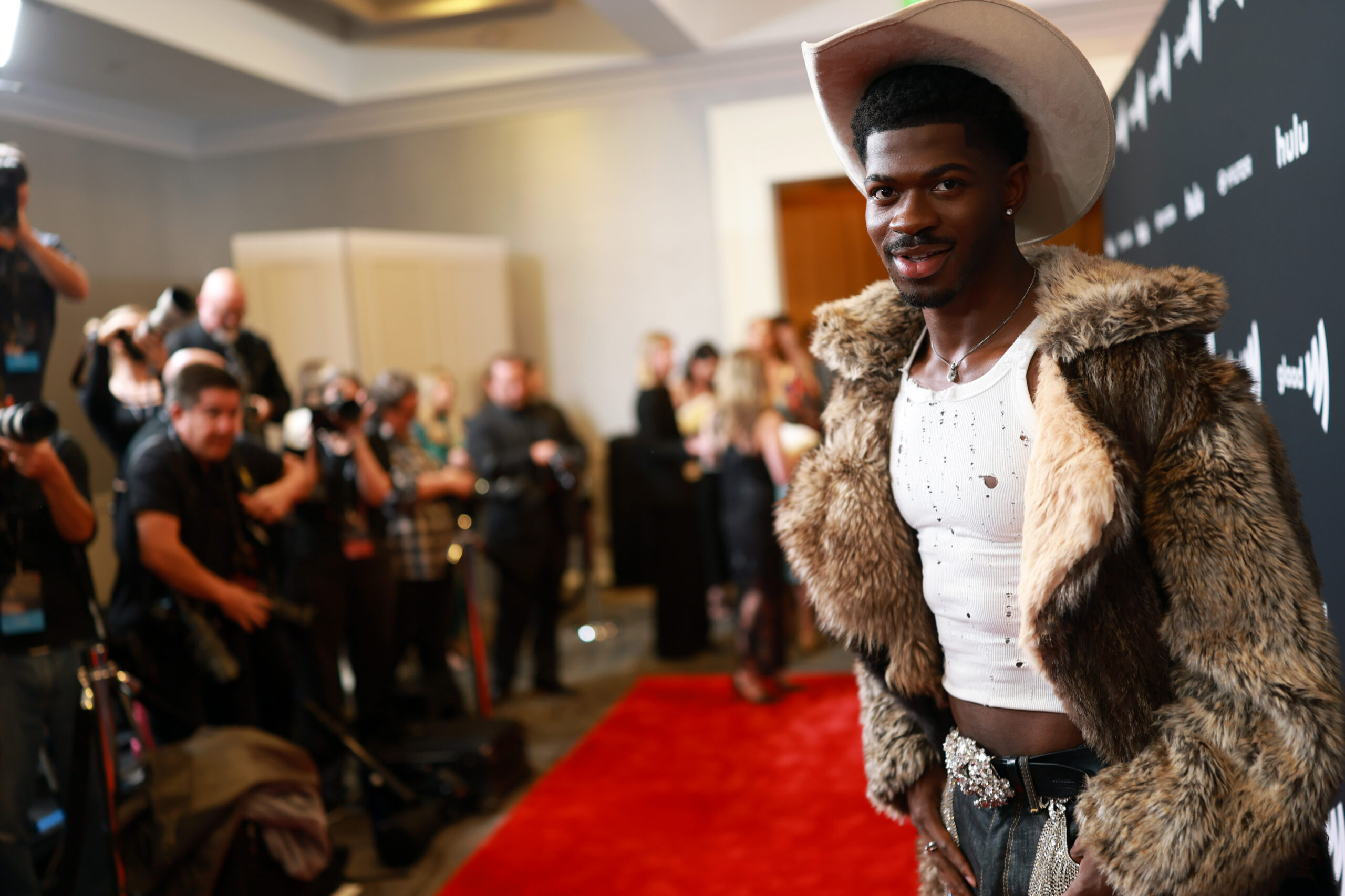 Male model on a red carpet posing for photographers, wearing a fur coat and wide-brimmed hat with a white shirt beneath.