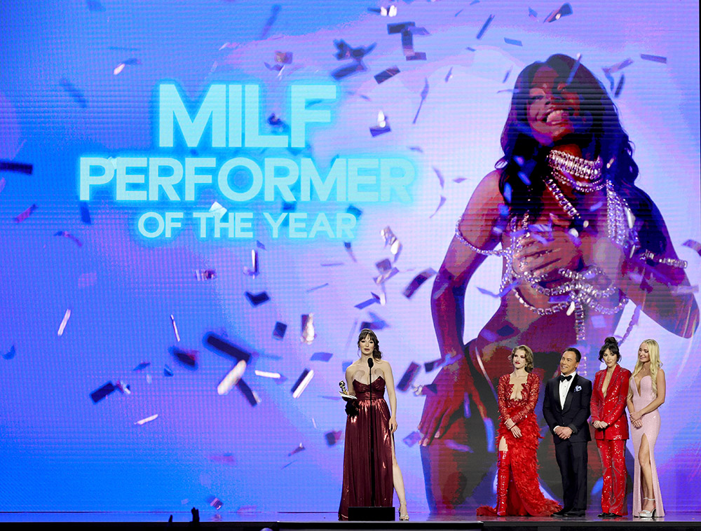 Award ceremony on a lit stage: five people in formal wear, confetti, and a large screen reading 'MILF PERFORMER OF THE YEAR'.