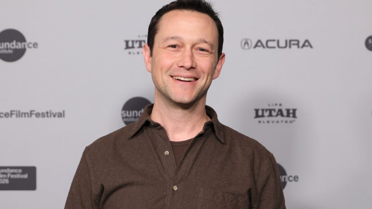 Smiling man in brown shirt posing in front of a sponsor backdrop at Sundance Film Festival.