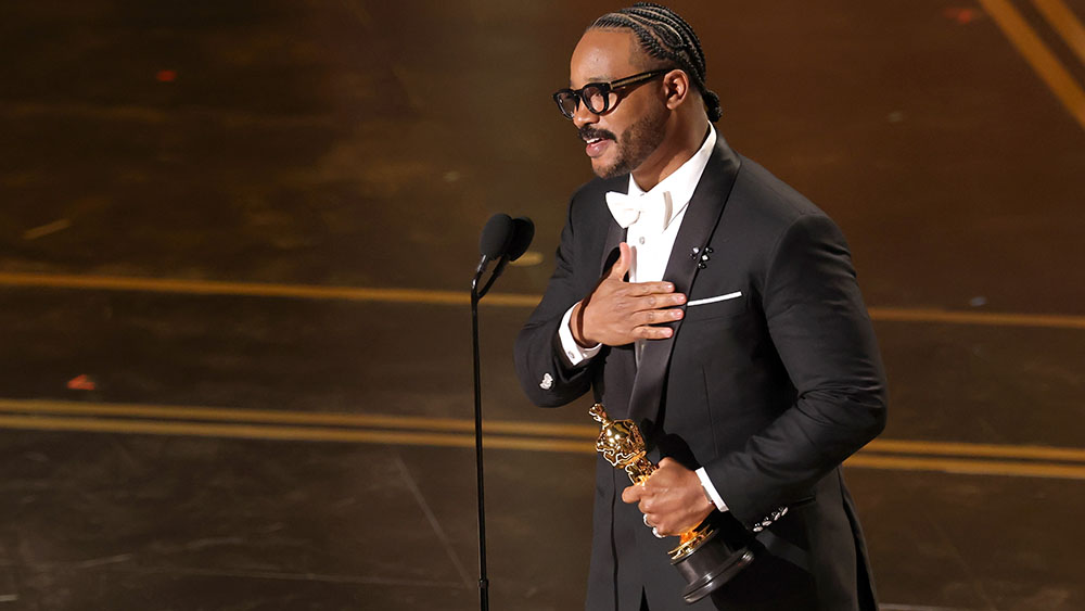 A man in a black tuxedo at a podium, holding an award and placing his hand over his heart on stage.